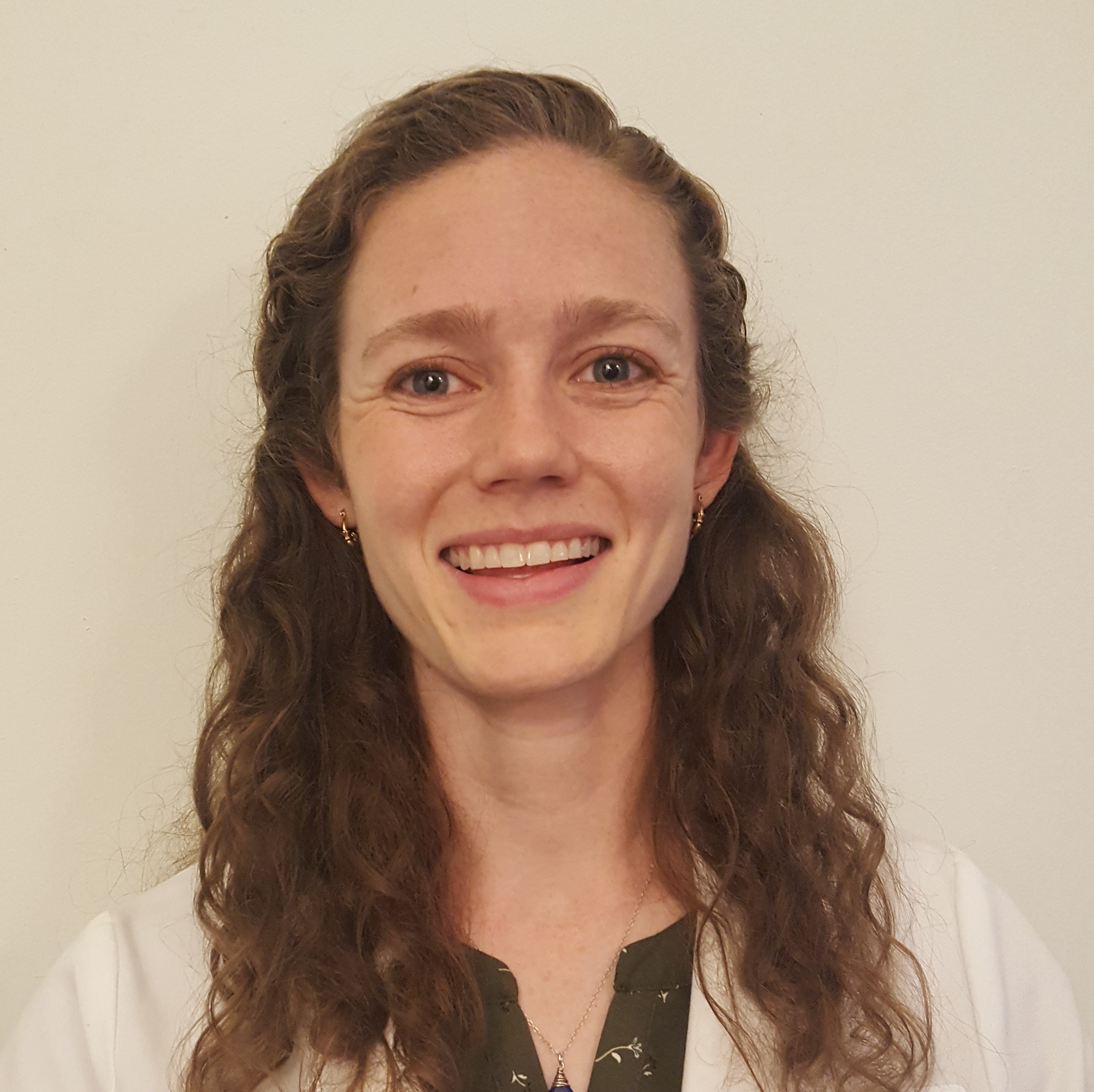 Alumna Rebecca Baldwin smiling, long light brown curly hair over her shoulders, wearing a white coat